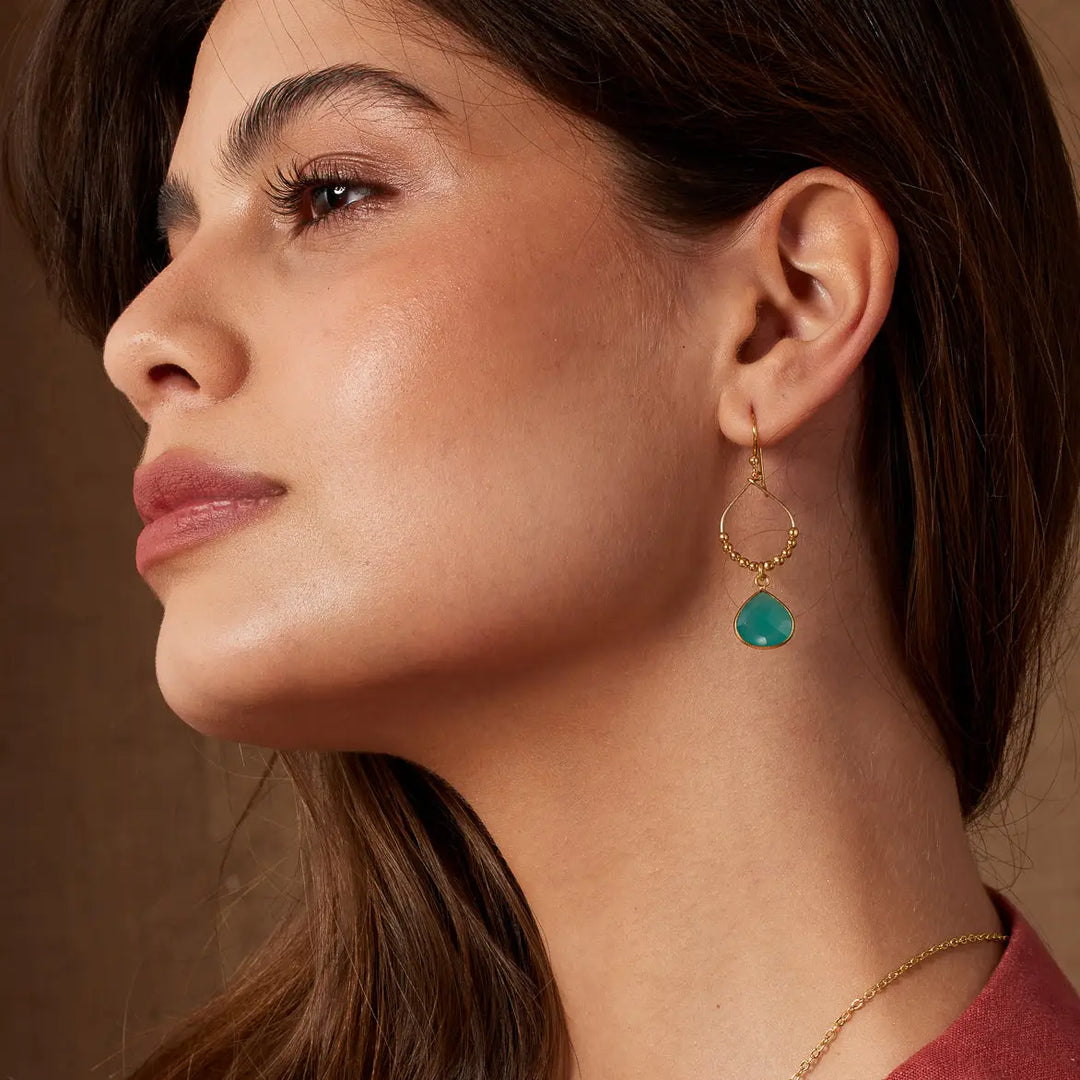 Woman with her head angled down. She is wearing gold earrings and 2 gold necklaces -one has a rectangular green-coloured pendant.