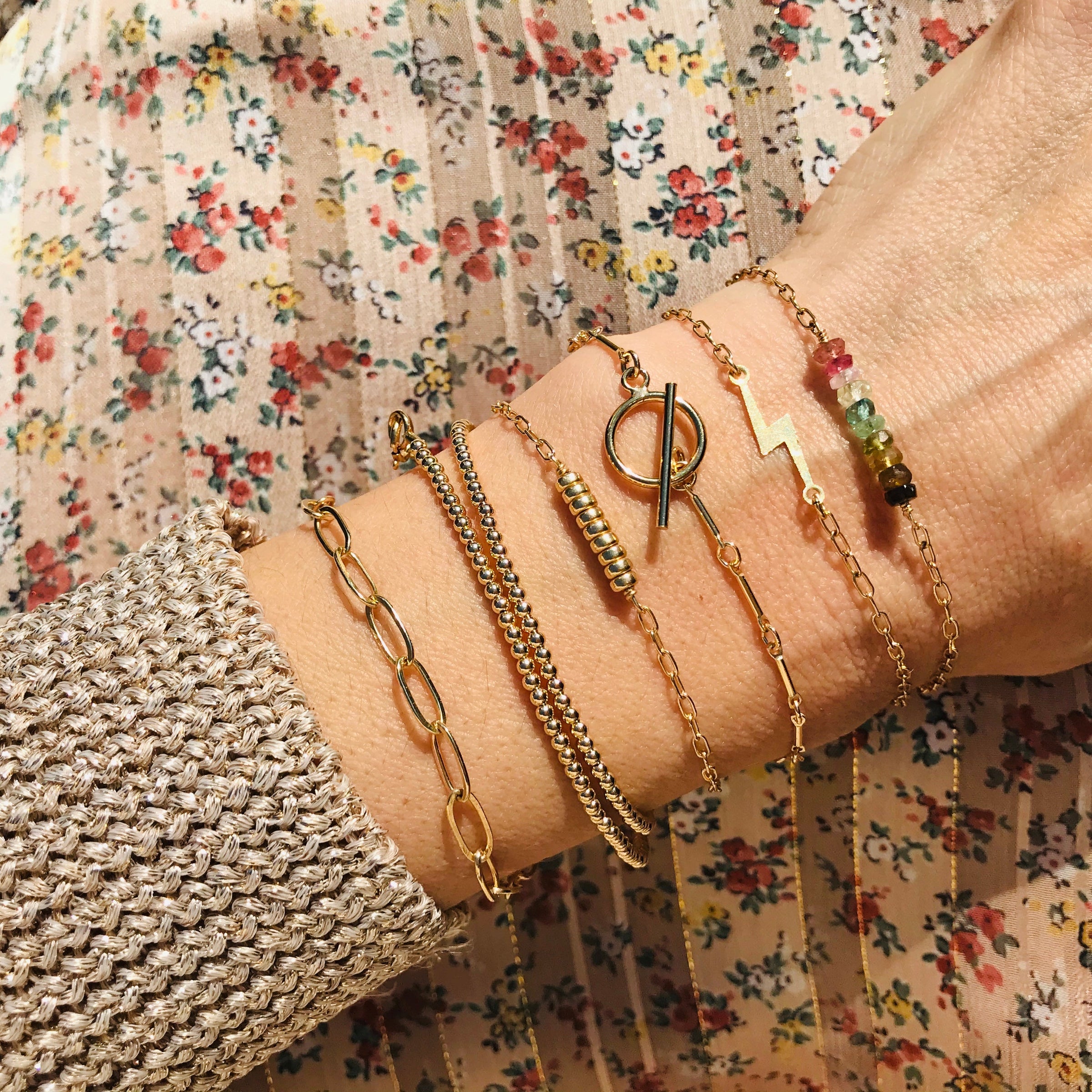 Woman's wrist with multiple gold-coloured bracelets. Her arm is resting on a beige & floral background.