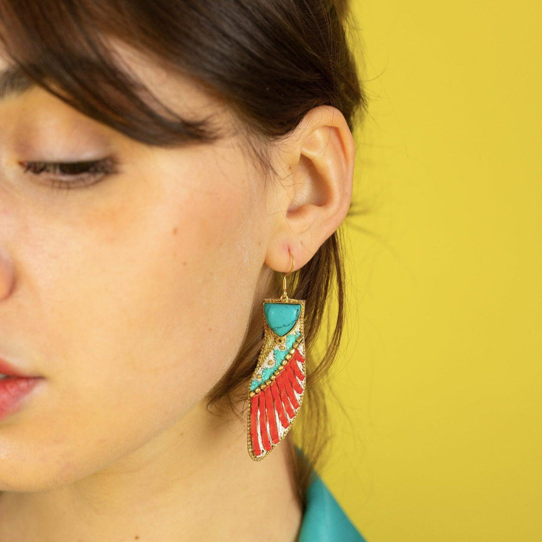A person wearing a turquoise top and showcasing a pair of earrings with multicolored sequins and brass beads against a yellow background.