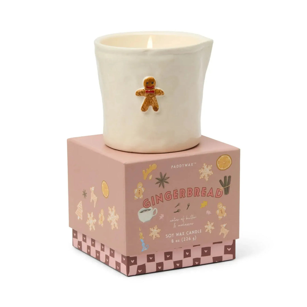 Candle in a decorative pot with a gingerbread man design, placed on a matching box with festive illustrations.