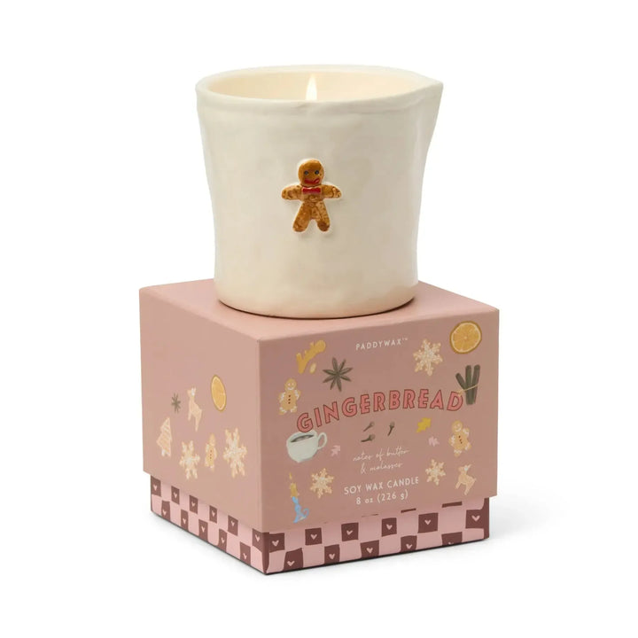 Candle in a decorative pot with a gingerbread man design, placed on a matching box with festive illustrations.