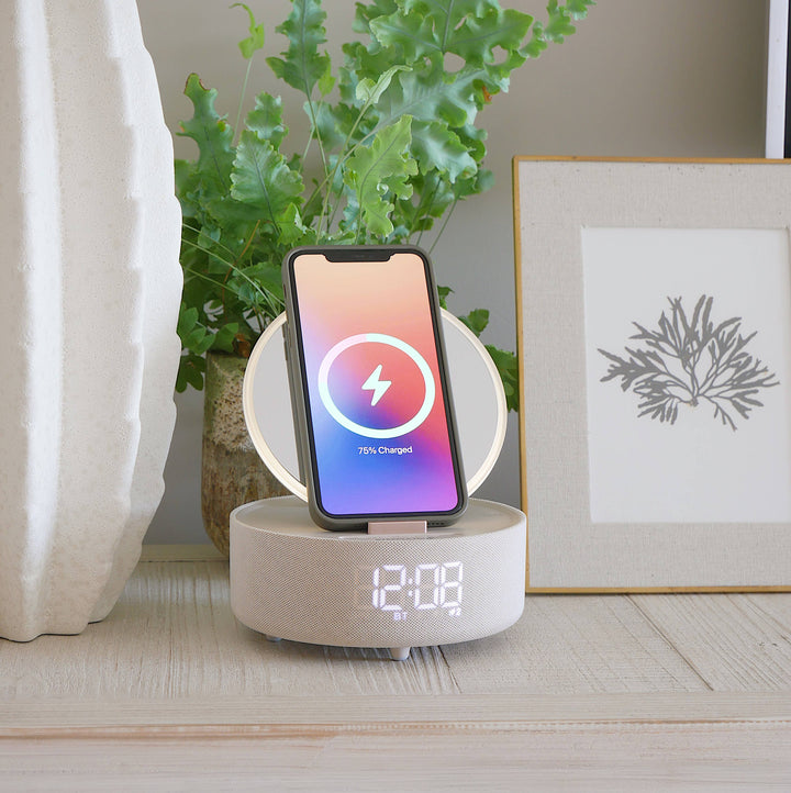 A multifunctional bedside device serving as a mirror, Bluetooth speaker, clock, and wireless charger, in a taupe color, placed on a table next to a plant and a framed picture.