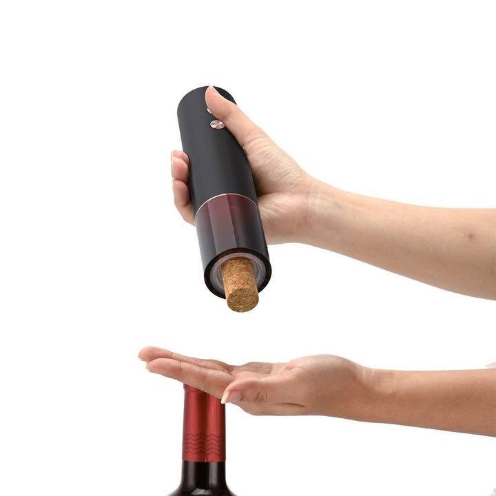 Hand using a black electric wine bottle opener to remove a cork from a red wine bottle on a white background.