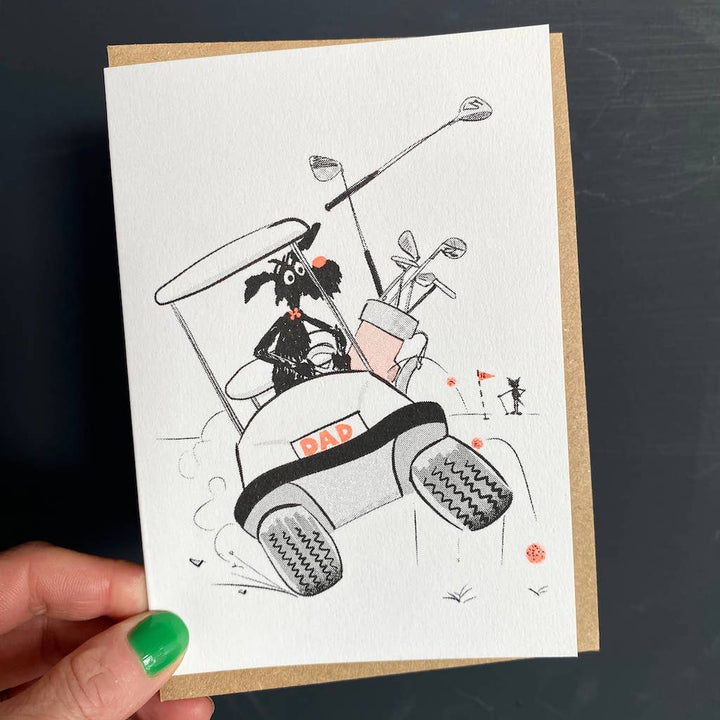 Greetings card with an illustration in black and white of a black dog driving a golf cart with the numberplate'DAD' in pale orange.