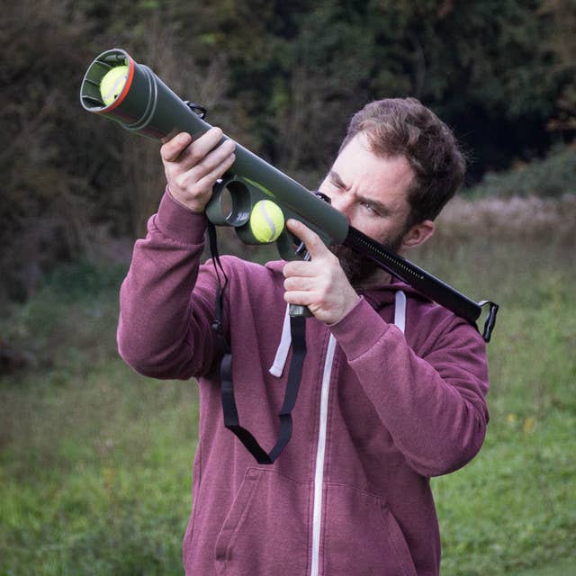Man holding a large green toy gun with tennis balls in a grassy field for dogs