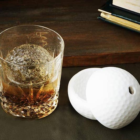 Golf ball-shaped ice cube tray next to a glass of whiskey with ice on a wooden surface.