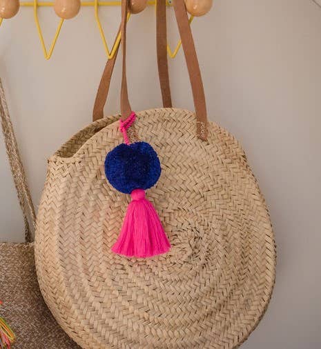A large indigo blue pom pom with a magenta pink tassel attached to a wicker handbag.