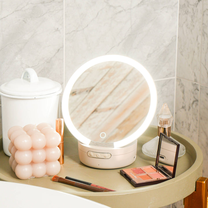Compact round makeup mirror with built-in Bluetooth speaker, displayed on a table alongside various makeup items like pens and a compact powder.
