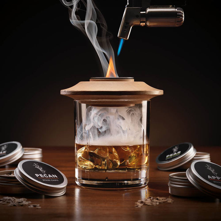 Glass container with liquid being heated by a torch, surrounded by flavor discs on a dark background. Smoke your whiskey