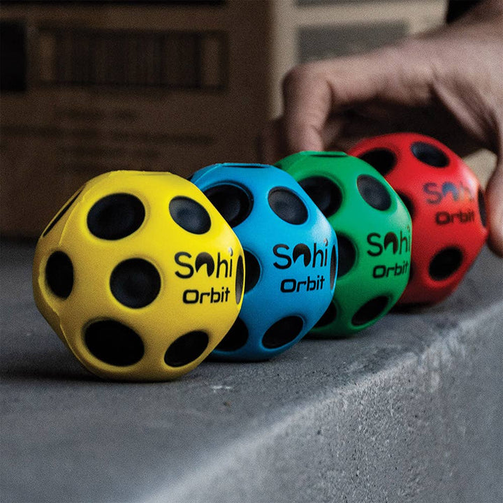 Four colorful 'Sahi Orbit' balls on a concrete surface with a hand reaching towards them.