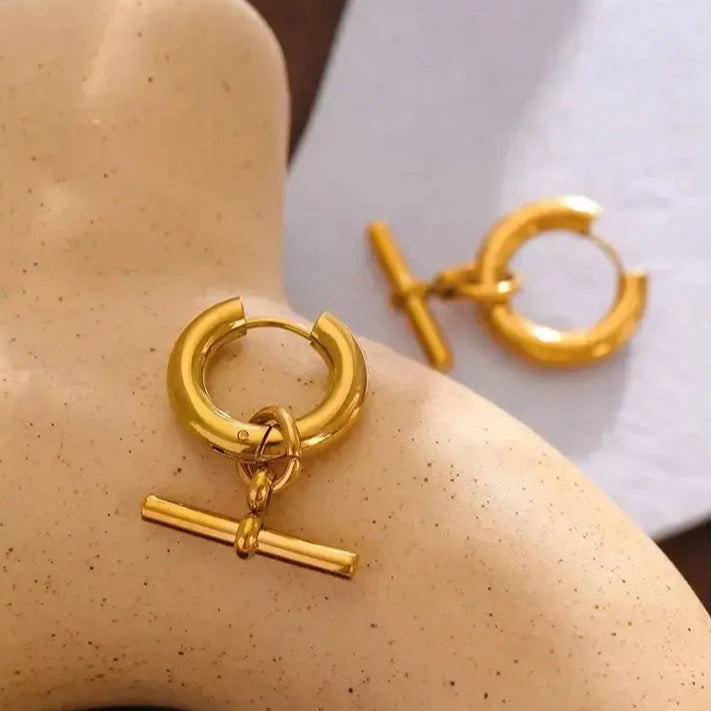 A pair of gold-plated hoop earrings with a T-bar detail displayed on a mannequin ear.