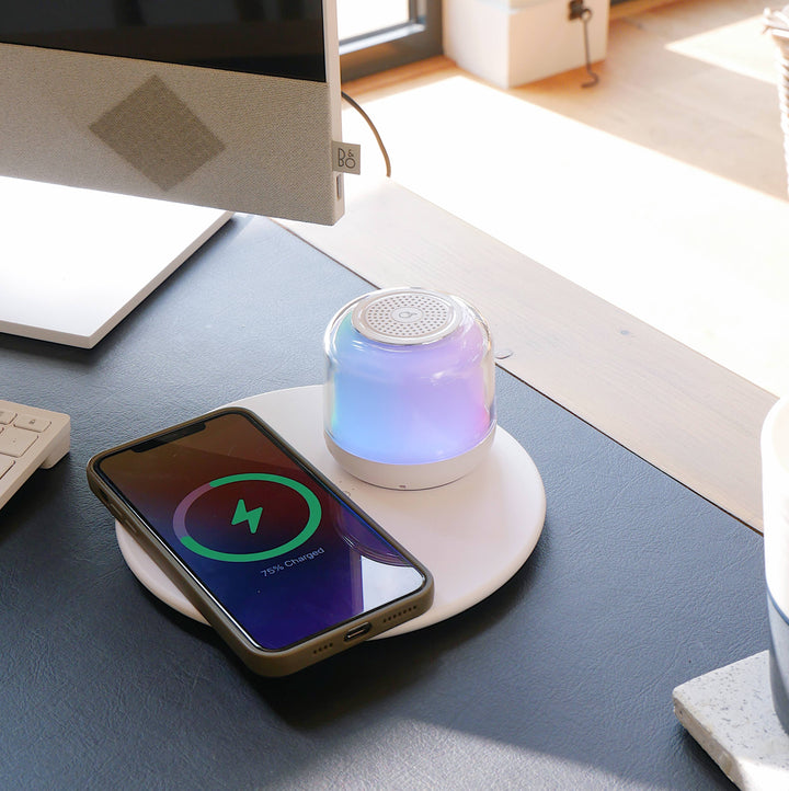 A wireless charger and Bluetooth speaker with a smartphone on top, displaying a colorful light base on a desk.