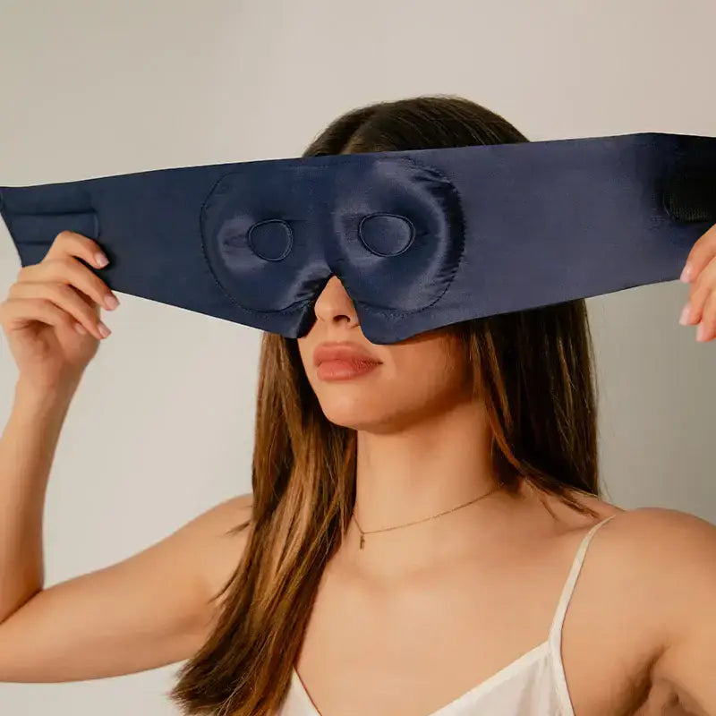 Woman holding a blue satin eye mask over her eyes against a plain background