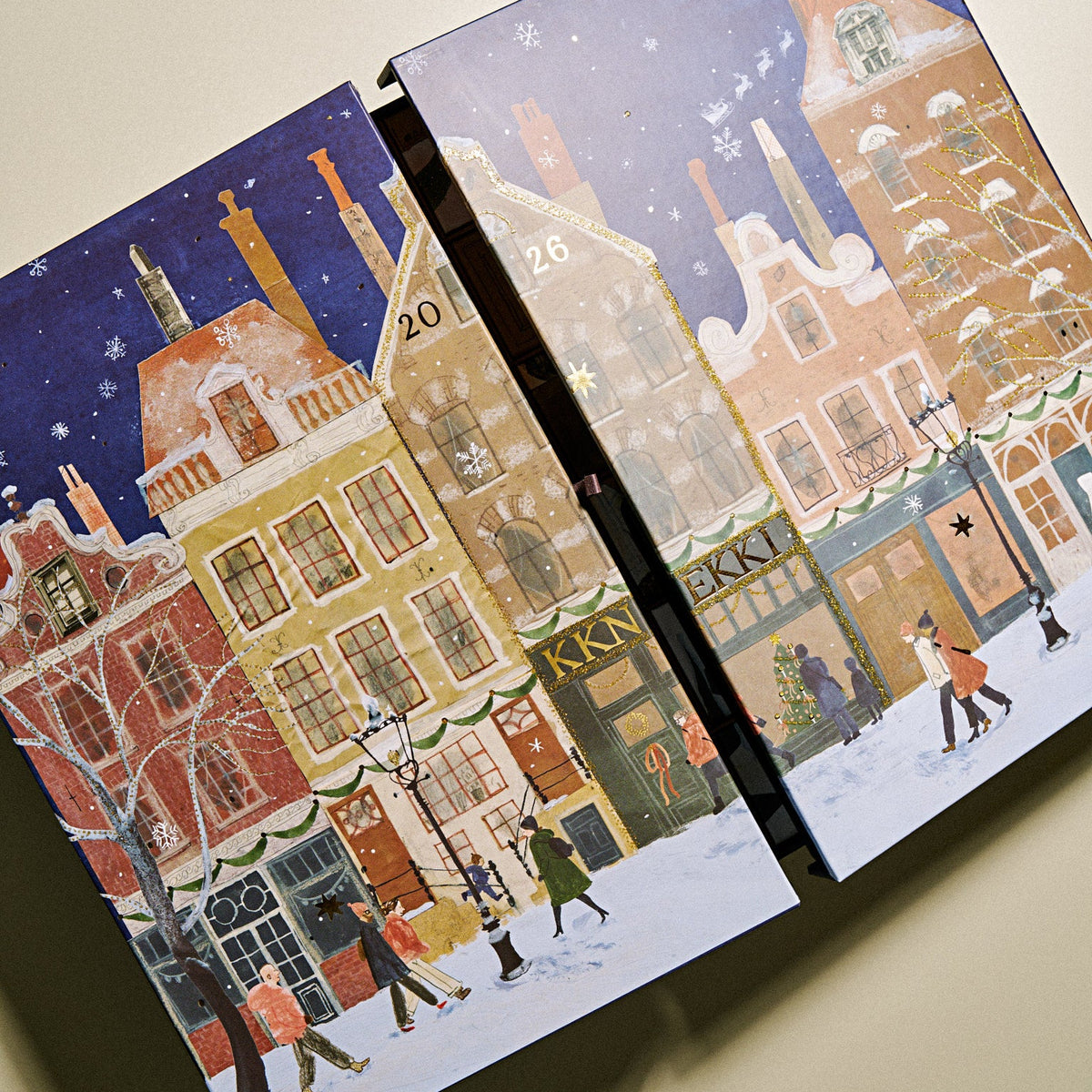 An KKNNEKKI illustrated advent calendar boxes shown at an angle against a plain beige background. The boxes feature a snowy winter street scene with colorful buildings, snow, stars, and small figures walking along the sidewalk.