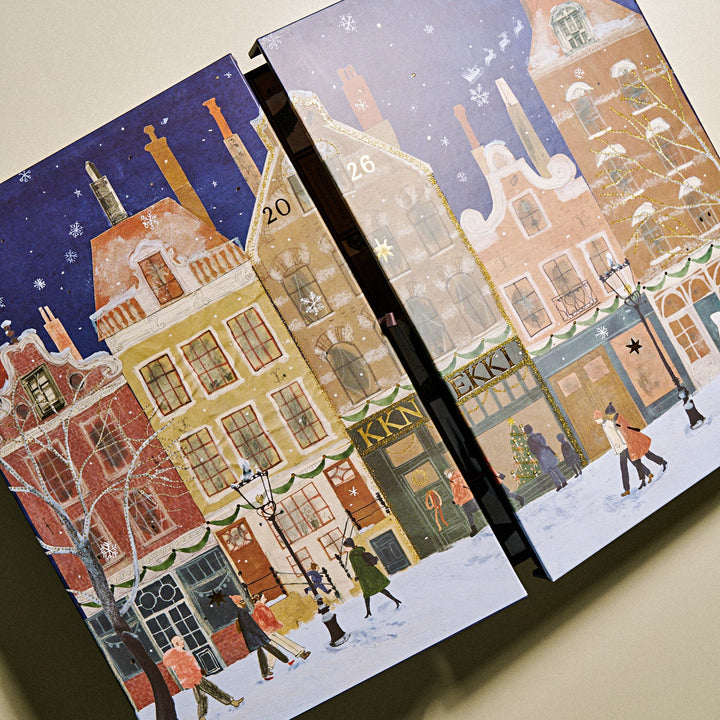 An KKNNEKKI illustrated advent calendar boxes shown at an angle against a plain beige background. The boxes feature a snowy winter street scene with colorful buildings, snow, stars, and small figures walking along the sidewalk.