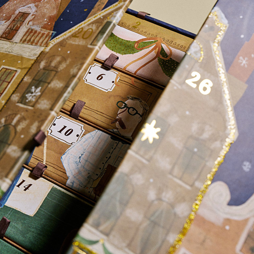 A close-up of a decorated KKNEKKI advent calendar box with illustrated holiday houses, numbered doors, and gold glitter trim. Visible numbers include 6, 10, 14, and 26.