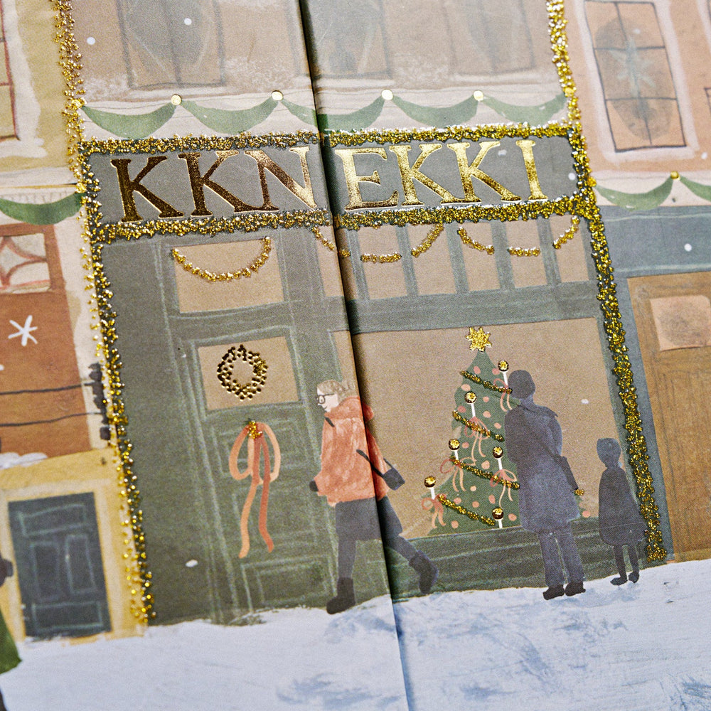 A close-up of the KKNEKKI 2026 Advent Calndar printed illustration showing a snowy storefront with the word 'KKN EKKI' in gold lettering, decorated with garlands and a Christmas tree, while people walk in front.
