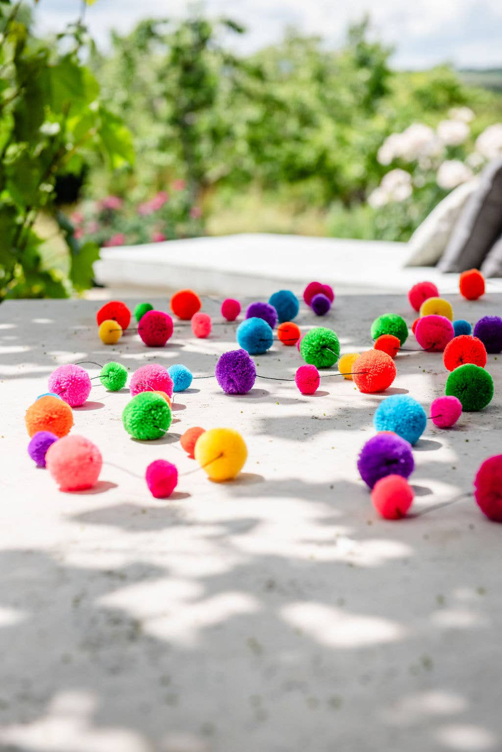 Brightly-coloured pom-pom garland on stone slabs with garden plants in the background.