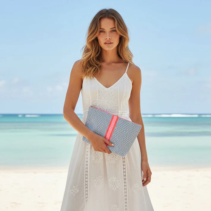 Woman holding a clutch bag on a beach with clear blue sky and ocean.