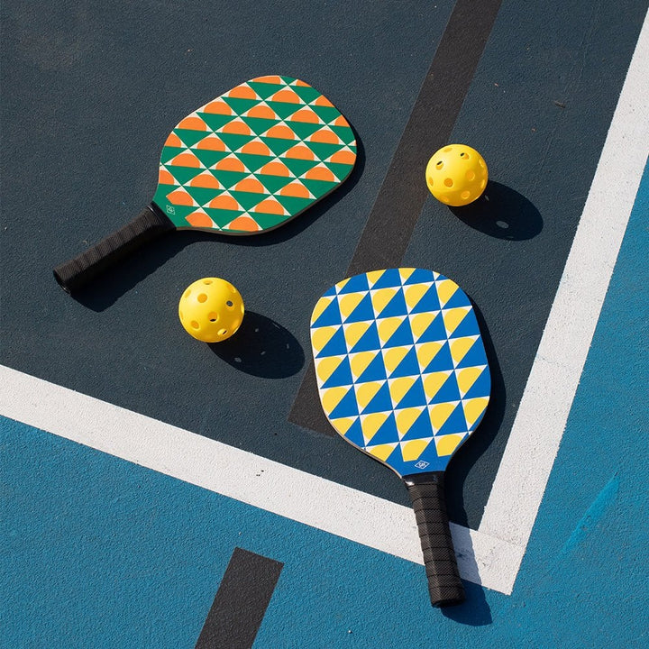 Two pickleball paddles with geometric patterns and two balls on a pickleball court.