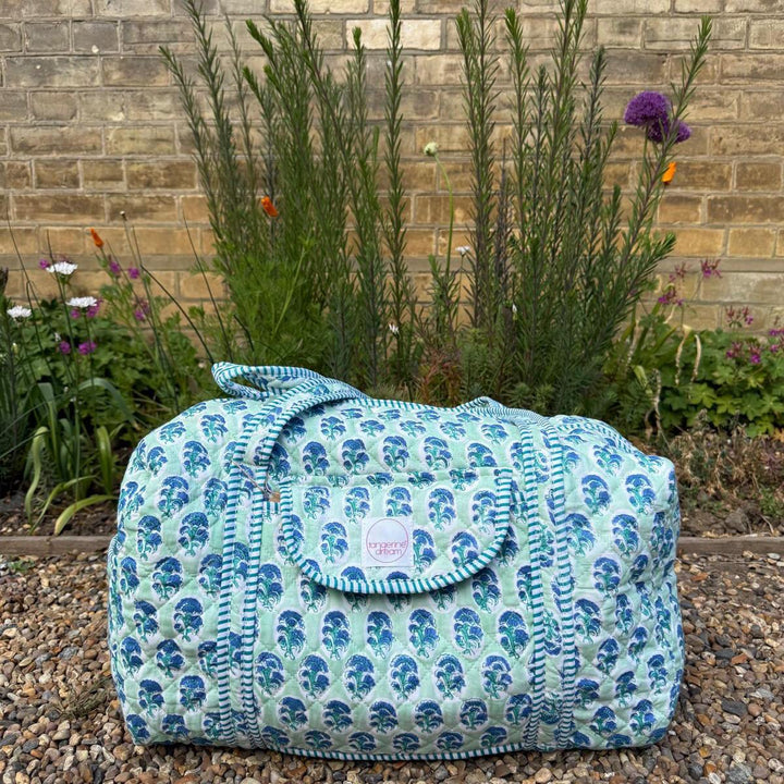 Blue floral-patterned bag on a gravel surface with a brick wall and plants in the background