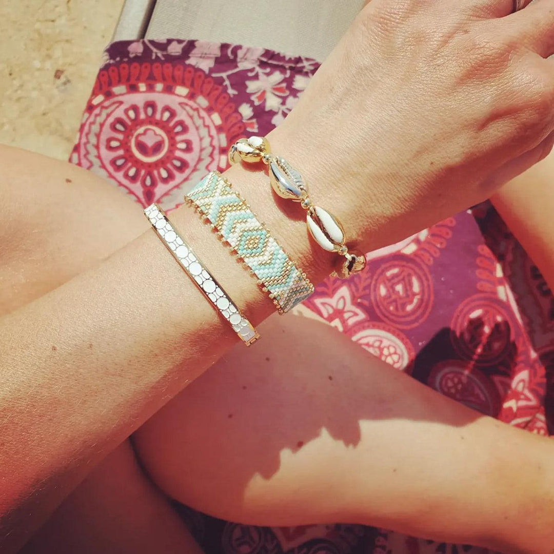 A person's wrist wearing a gold bracelet with white enamel details, displayed against a patterned background.