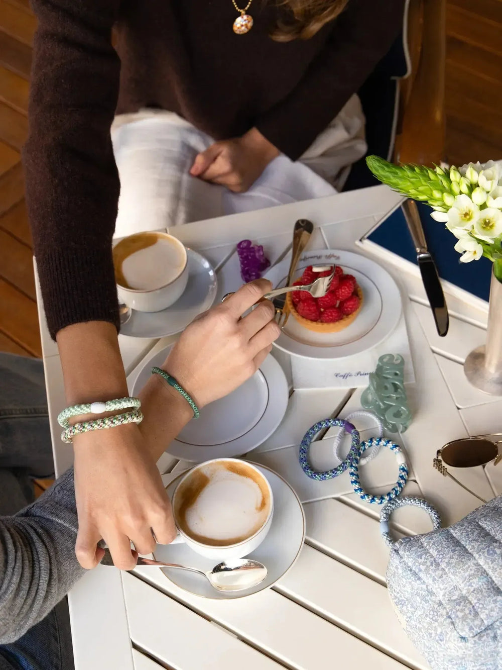Looking down at a white table with coffee cups, a fruit tart and two arms with hairbands on wrists.