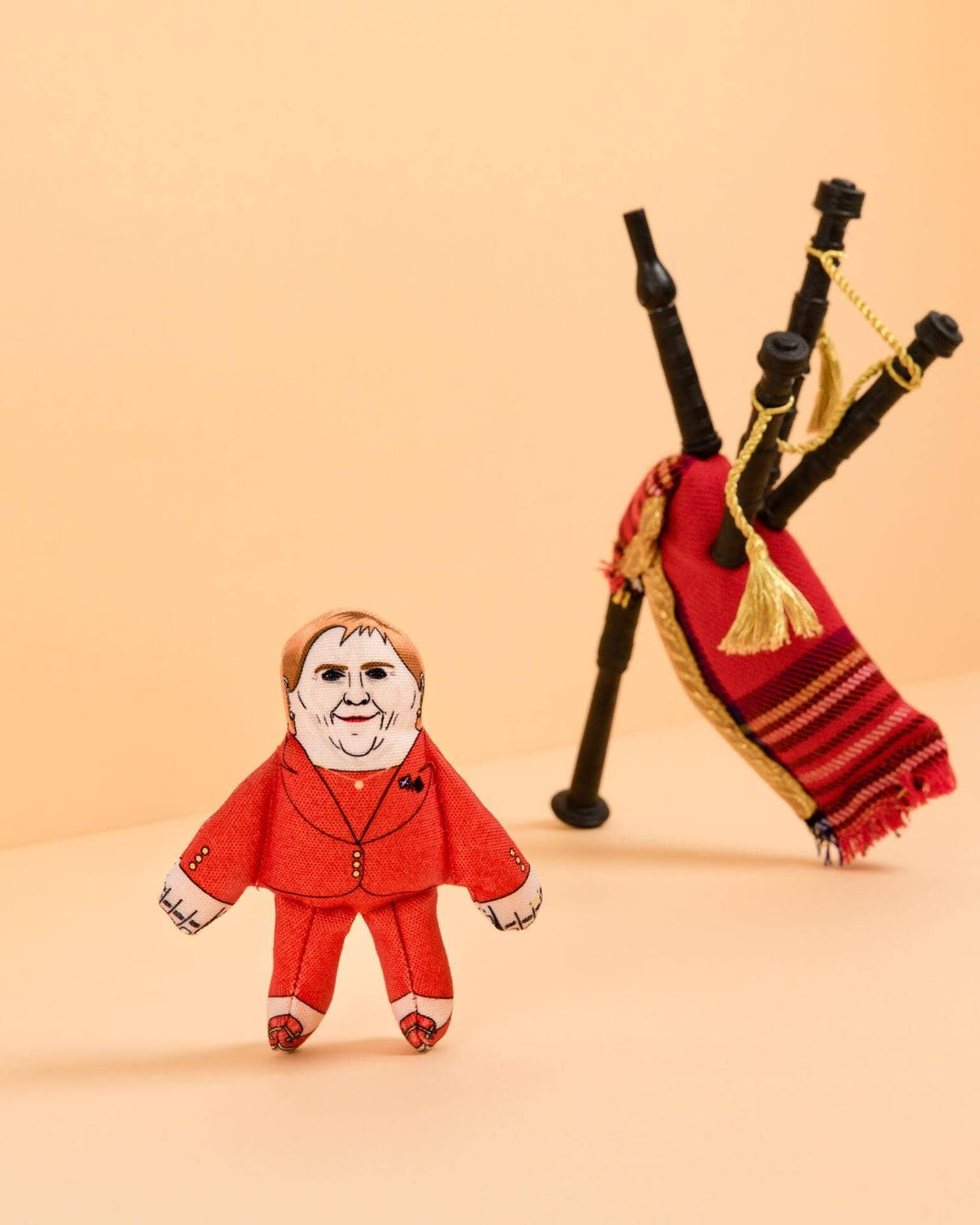 Small plush toy figure in a red outfit next to bagpipes on a beige background