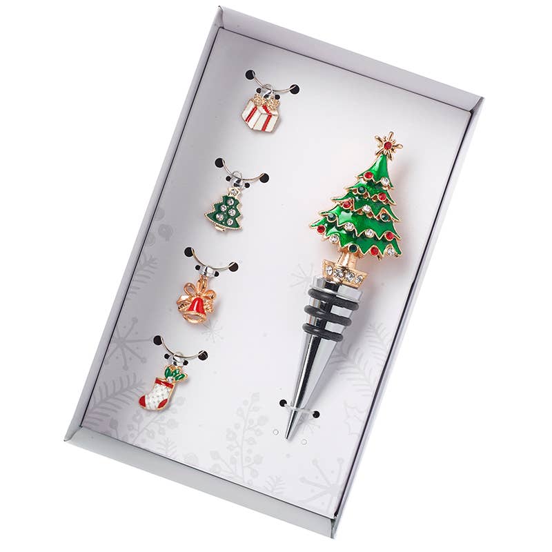 A Christmas-themed bottle stopper set displayed inside a white gift box. The set includes a main bottle stopper shaped like a decorated Christmas tree with gold-tone metal, green enamel, red accents, and a small gold bow on top, plus four matching small holiday charms (gift box, Christmas tree, bell, stocking) arranged beside it.